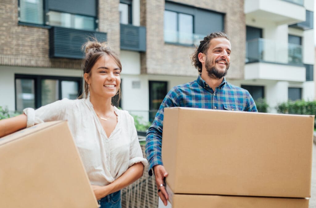 Young multifamily residents carrying moving boxes into an apartment building during summer move-in season, highlighting the need for secure package delivery solutions like the Smart Package Room system