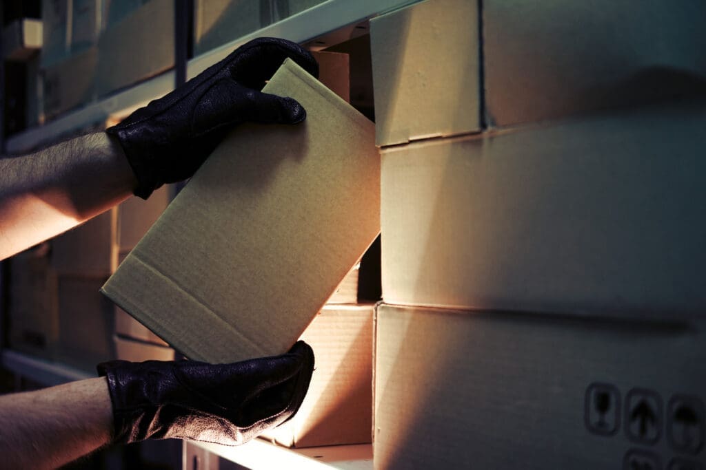 Person wearing black gloves removing a package from a storage shelf, illustrating the risk of package theft in unsecured delivery areas.